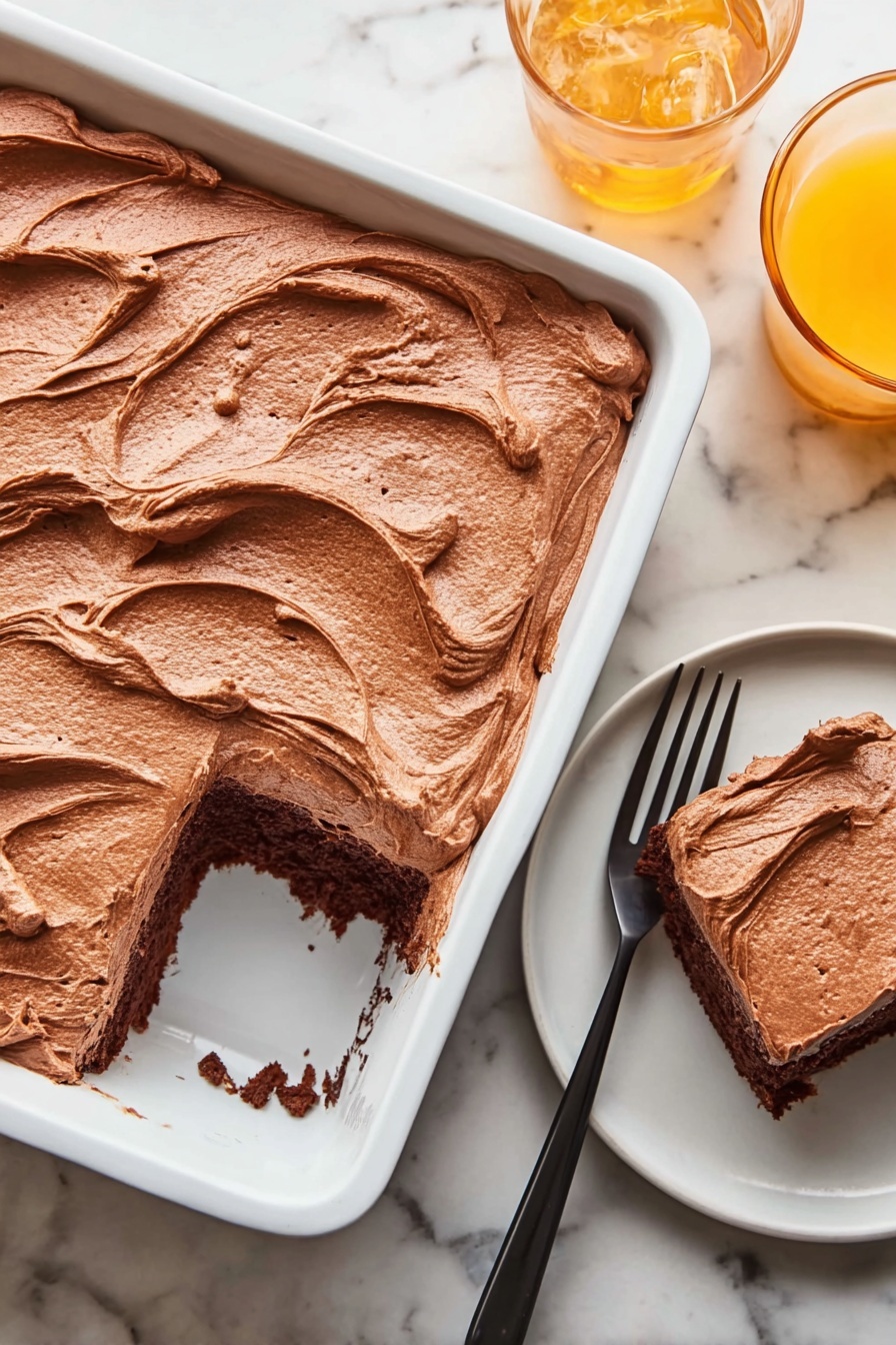 A white rectangular baking dish holds a thick layer of smooth, light brown chocolate frosting spread evenly, with visible swirls and soft peaks on top. One square piece has been removed from the bottom right corner of the dish, showing a dark brown chocolate cake layer beneath the frosting. Next to the dish, on a small white round plate, sits the removed cake piece with the same rich frosting on top, accompanied by a black fork resting beside it. In the top right corner, there is a glass of orange drink on the white marbled surface. Photo taken with an iphone --ar 2:3 --v 7 - Chocolate Mayonnaise Sheet Cake, moist chocolate cake with mayonnaise, easy chocolate cake recipe, homemade chocolate sheet cake, rich chocolate dessert