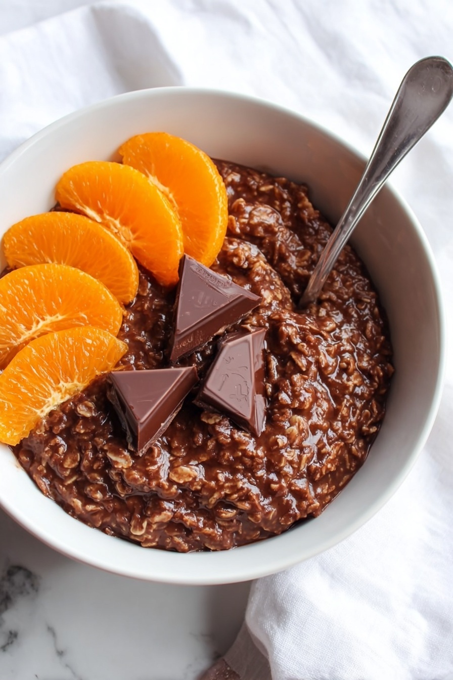 The image shows a white bowl filled with a thick, dark brown chocolate oatmeal mixture that has a slightly rough texture from the oats. On the top left side of the oatmeal, there are six orange slices arranged in a neat cluster, while on the top right side, two triangular pieces of rich, dark chocolate rest partially melted on the oatmeal's surface. A silver spoon is placed inside the bowl on the right side, angled upward. The bowl sits on a white marbled surface with part of a white cloth visible in the background. Photo taken with an iphone --ar 2:3 --v 7 - Chocolate Orange Porridge, breakfast ideas with orange and chocolate, easy healthy porridge recipes, cozy morning breakfast, citrus chocolate oatmeal
