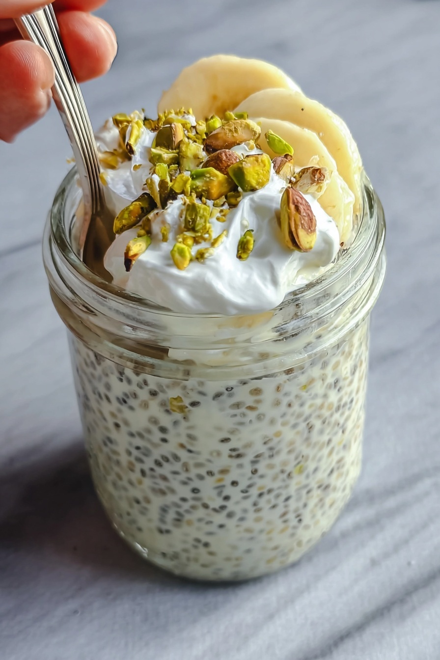 The image shows a clear glass jar filled almost to the top with a creamy white chia pudding that has small black chia seeds spread evenly throughout. On the top layer inside the jar, there are a few slices of light yellow banana, which are partially covered by the creamy pudding. Over the bananas, there is a layer of small white whipped cream dollops, adding a soft texture. Scattered on the very top are roughly chopped green pistachio nuts that add a contrasting color and crunchy texture. A spoon is placed inside the jar, with a woman's hand holding it gently from the left side. The jar is set against a smooth, white marbled surface. Photo taken with an iphone --ar 2:3 --v 7 - High Protein Pistachio Overnight Oats, healthy overnight oats, protein breakfast recipes, pistachio flavored oatmeal, make-ahead breakfast ideas