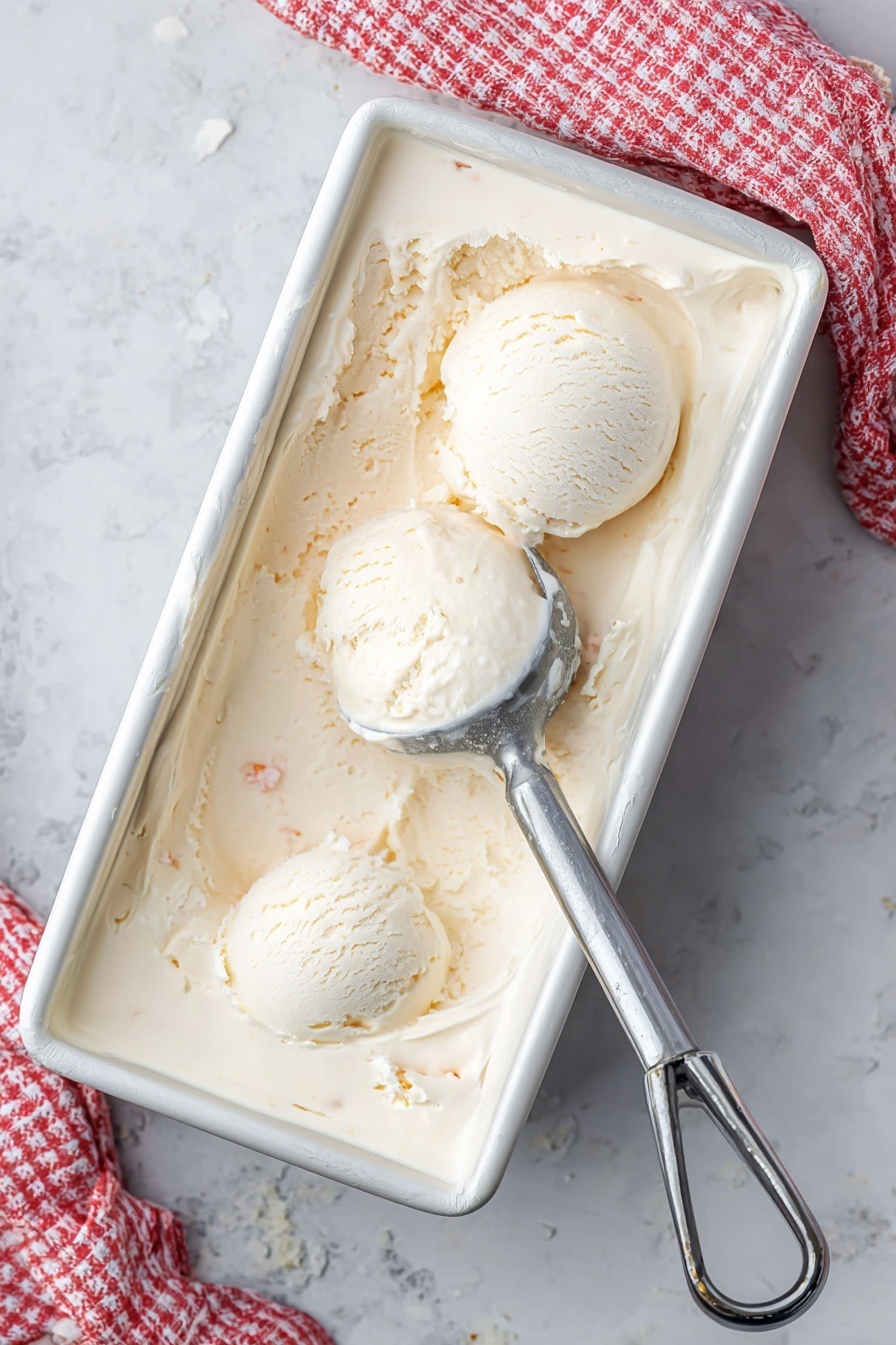 A white metal rectangular container filled with creamy, pale beige ice cream shows two round scoops placed near the top left corner. Ice cream has a smooth but slightly textured surface with small visible bits inside. A silver ice cream scooper rests partially buried on the right side, with some ice cream residue on it. A white and red checkered cloth is partly visible in the upper right corner on a white marbled surface. photo taken with an iphone --ar 2:3 --v 7 - Homemade Coconut Ice Cream, Coconut Ice Cream, Tropical Coconut Dessert, No-Churn Coconut Ice Cream, Dairy-Free Coconut Ice Cream
